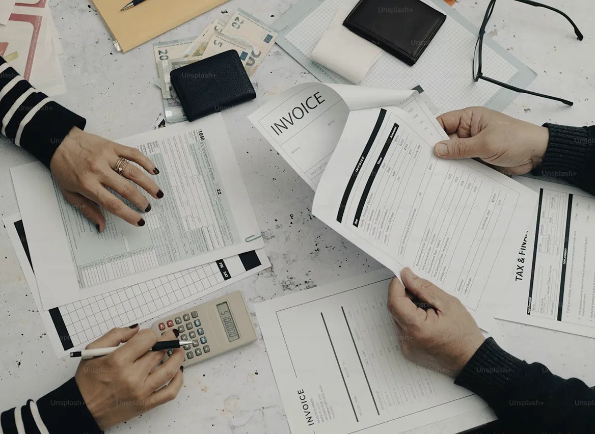 Twee personen bekijken facturen en formulieren op een bureau met een rekenmachine, portemonnees en eurobiljetten eromheen.